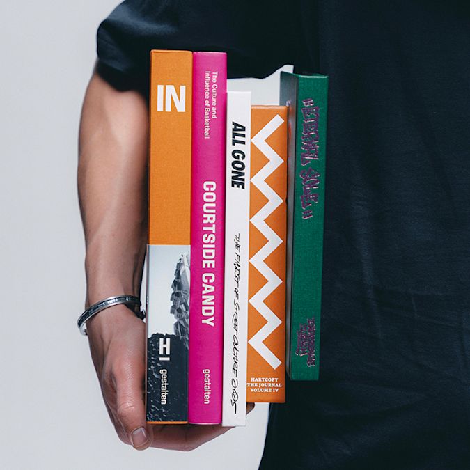 Person holding a stack of colorful books with visible titles and graphic spines against a neutral background, wearing a black short-sleeve shirt and a bracelet.