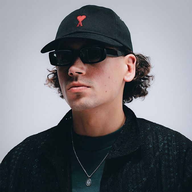 Close-up of a person wearing a black baseball cap with a red heart logo, black rectangular sunglasses, and a textured black jacket, set against a light background.