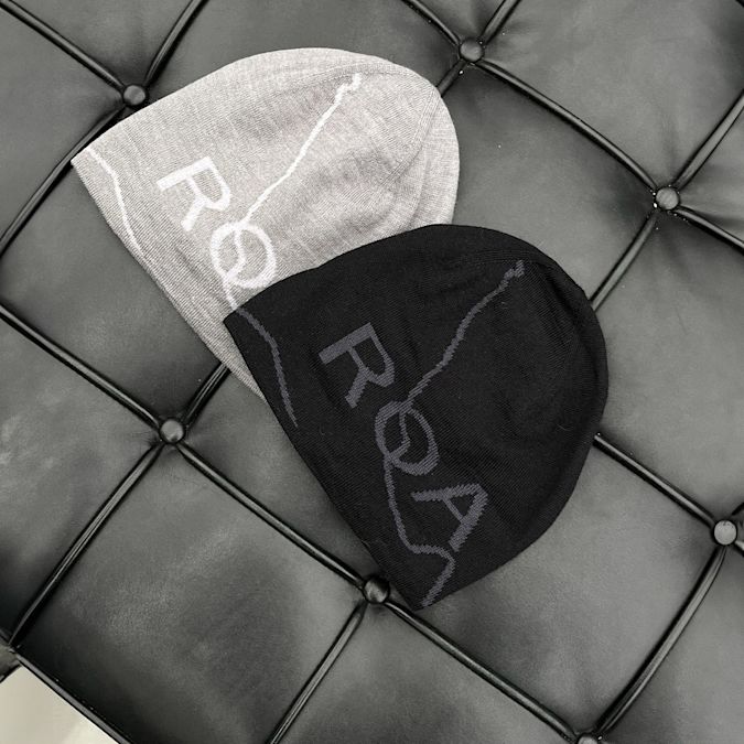 Two beanies, one gray and one black, each with the word “ROA” and a wavy line design, placed on a tufted black leather surface.