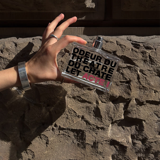A hand with silver rings and bracelet holds a perfume bottle labeled “ODEUR DU THEATRE DU CHÂTELET, ACTE I” in bold black and pink letters, against a sunlit stone wall.