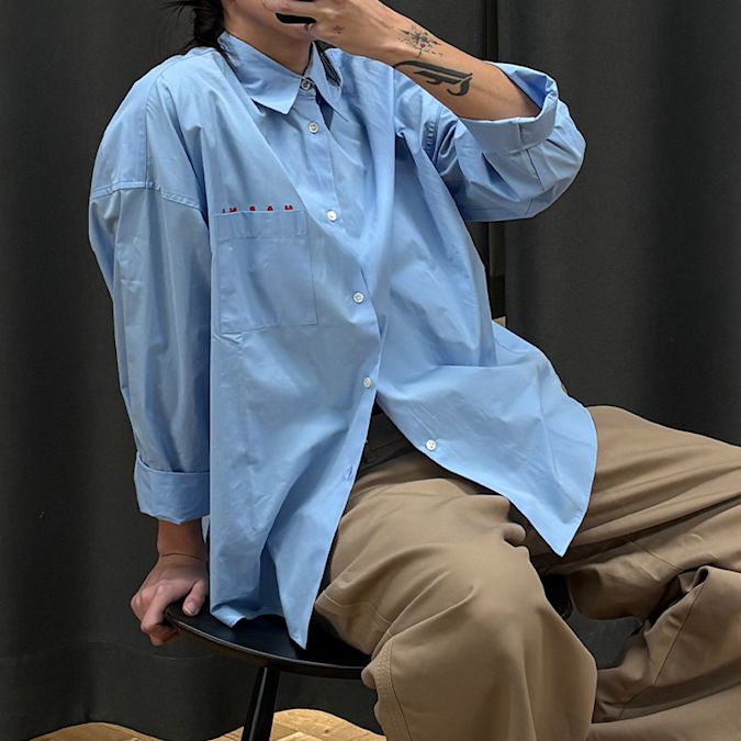 Person sitting on a stool wearing an oversized light blue button-up shirt with rolled sleeves and beige trousers, photographed against a dark curtain background.