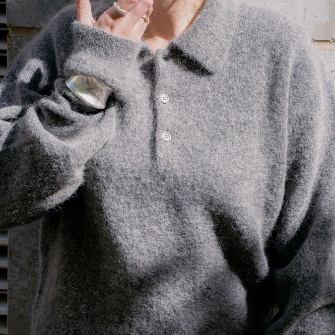 This image shows a close-up of a soft, fuzzy grey knit pullover with a polo-style collar and two white buttons. The knitwear has a brushed or mohair-like texture, giving it a cozy, slightly fluffy appearance. The wearer is accessorized with chunky silver rings and a bold silver cuff bracelet, adding a modern, edgy contrast to the softness of the sweater.
