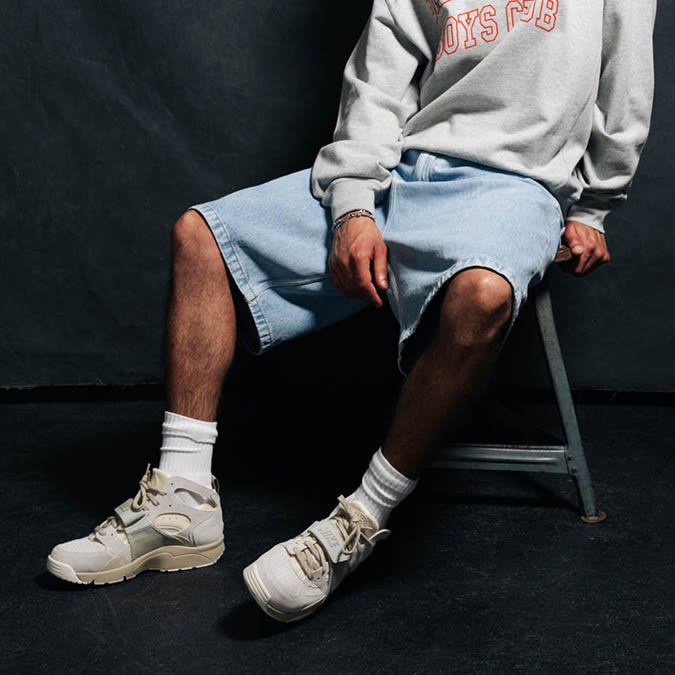 A man wearing a grey sweatshirt with red text, light blue baggy denim shorts, white socks, and beige chunky sneakers, sitting on a metal stool in a studio setting.