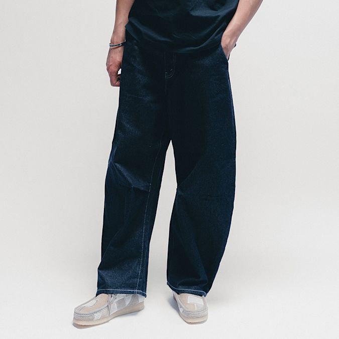 Lower-body studio shot of a person wearing loose, dark blue wide-leg jeans paired with light-colored sneakers, standing against a plain light background with one hand in a pocket.
