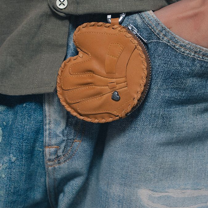 Close-up of a small brown leather coin pouch with stitched detailing, attached to the waistband of light blue washed jeans and partially tucked beneath a dark button-up shirt.