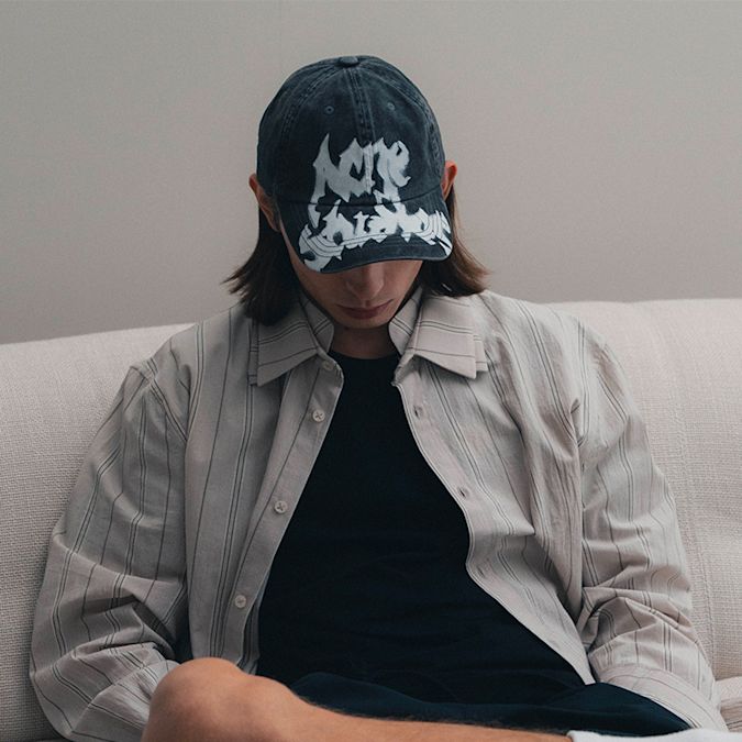 Close-up of a person wearing a black graphic baseball cap with a light striped button-up shirt layered over a black top, seated on a light-colored sofa.  Geben Sie Ihr Feedback zu BizChat ab.