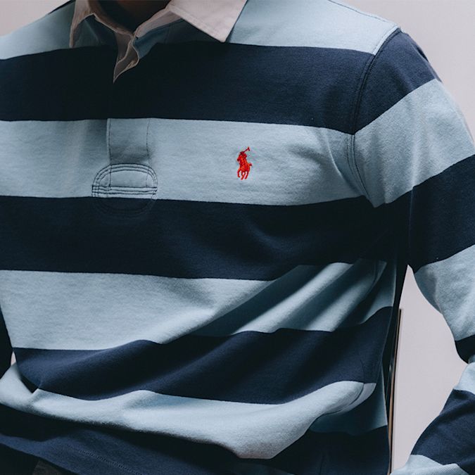 Close-up of a blue and navy horizontally striped long-sleeve rugby shirt with a white collar and a red embroidered Polo Ralph Lauren logo on the chest.