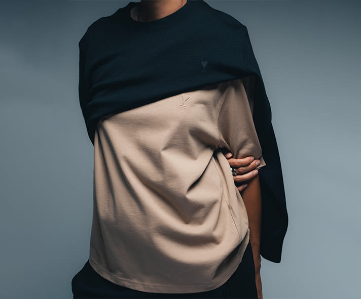 AMI Paris oversized beige T-shirt layered with a dark navy sweater draped over the shoulders, featuring subtle logo embroidery and a minimalist studio setting.
