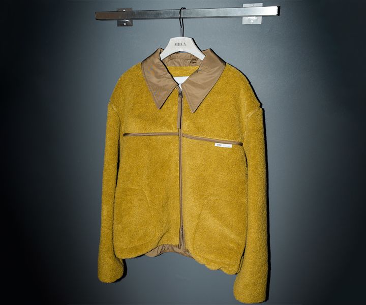 The image shows a mustard yellow fleece jacket hanging on a hanger labeled MBCY. The jacket features a prominent brown contrasting collar and zipper closure down the front. The material appears to be textured, likely sherpa or teddy fleece, giving it a cozy look. There’s a visible label near the chest zipper that reads “ami”, indicating it is from the French fashion brand AMI Paris.