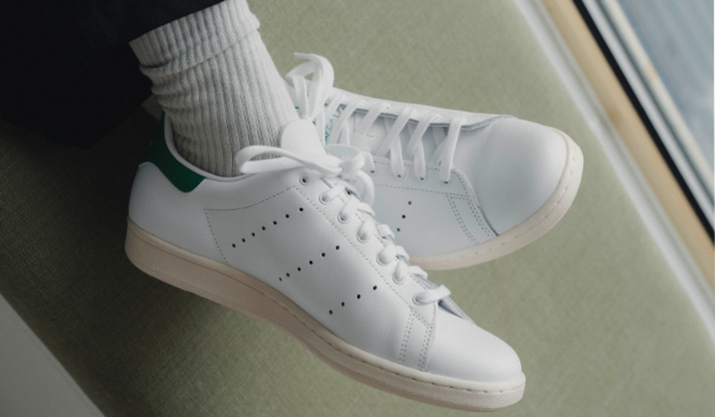 White adidas Stan Smith sneakers with green heel tabs worn with white ribbed socks, photographed from above on a light green surface.