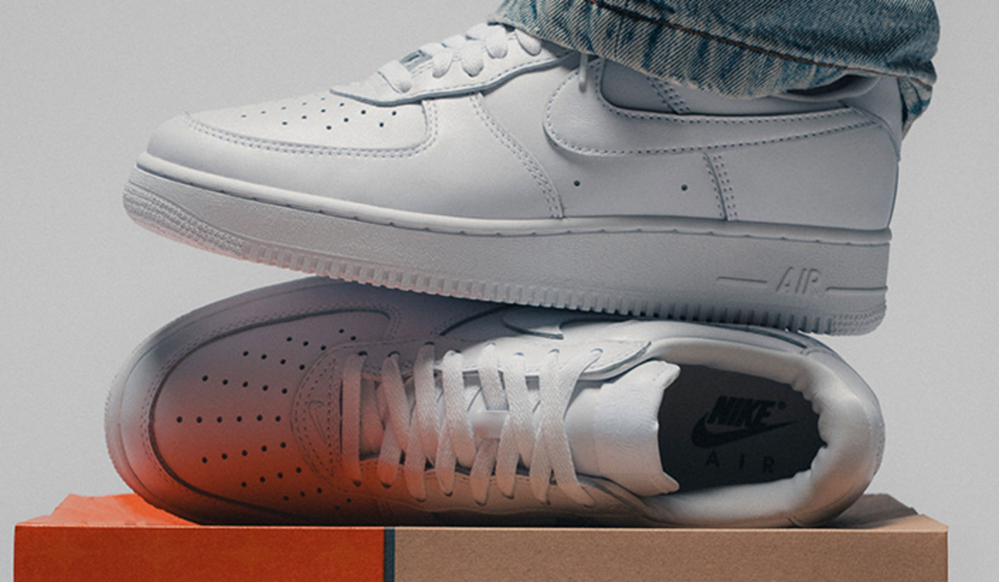 Close-up of a white Nike Air Force 1 ’07 sneaker worn with blue denim jeans, displayed above a second matching sneaker resting on stacked Nike shoeboxes against a clean studio background.