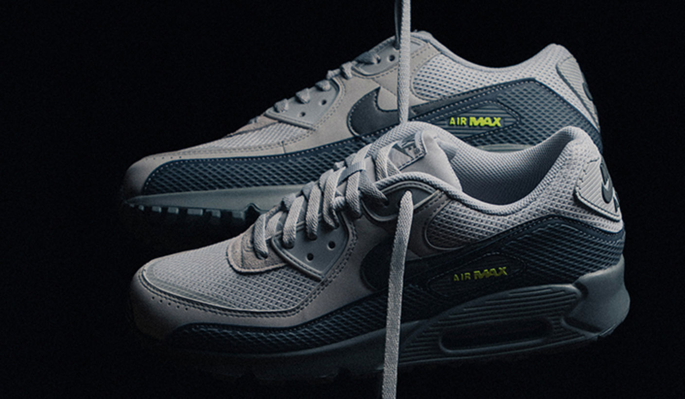Nike Air Max 90 “Grey Fog” sneakers in light and dark grey tones, shown in a low-light studio shot with neon green Air Max branding.