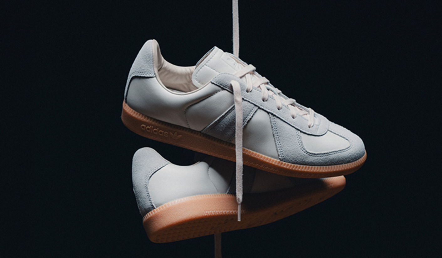 Pair of adidas BW Army sneakers in white leather with suede overlays and gum soles, suspended against a dark background with laces hanging down.