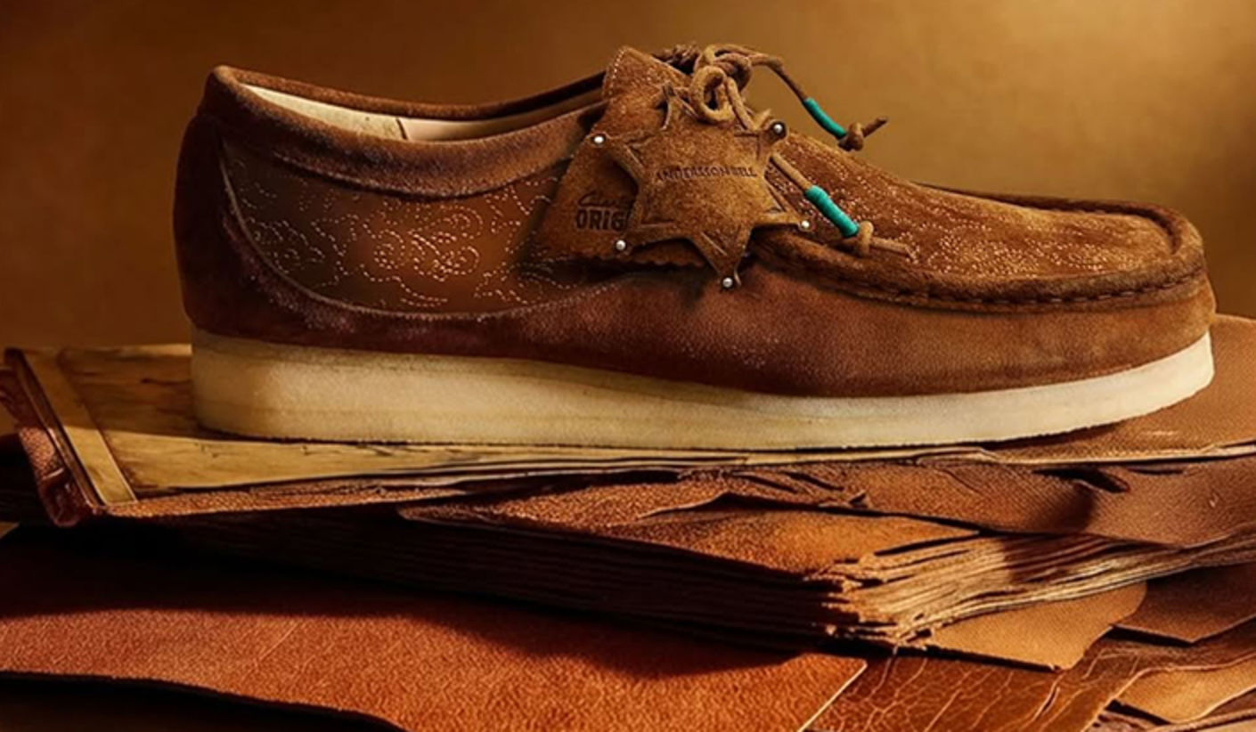 Clarks x Andersson Bell Wallabee shoe in brown suede with embossed detailing and decorative tags, placed on stacked leather materials against a warm-toned background.