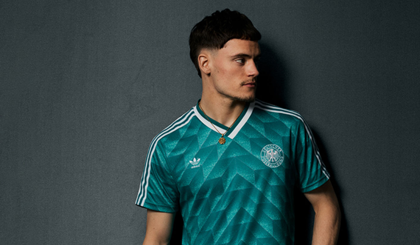 Florian Wirtz wearing a teal adidas DFB Germany 1990 away jersey featuring a geometric pattern, white shoulder stripes, V-neck collar, adidas logo, and German football federation crest, looking to the side against a plain grey background.