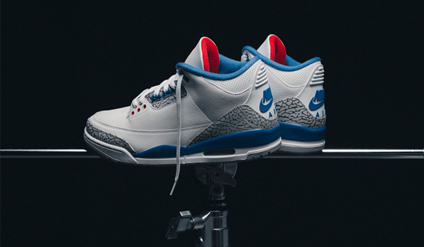 Pair of Air Jordan 3 Retro “True Blue” sneakers in white, blue, and red, displayed on a stand against a dark background, highlighting the iconic elephant print detailing.