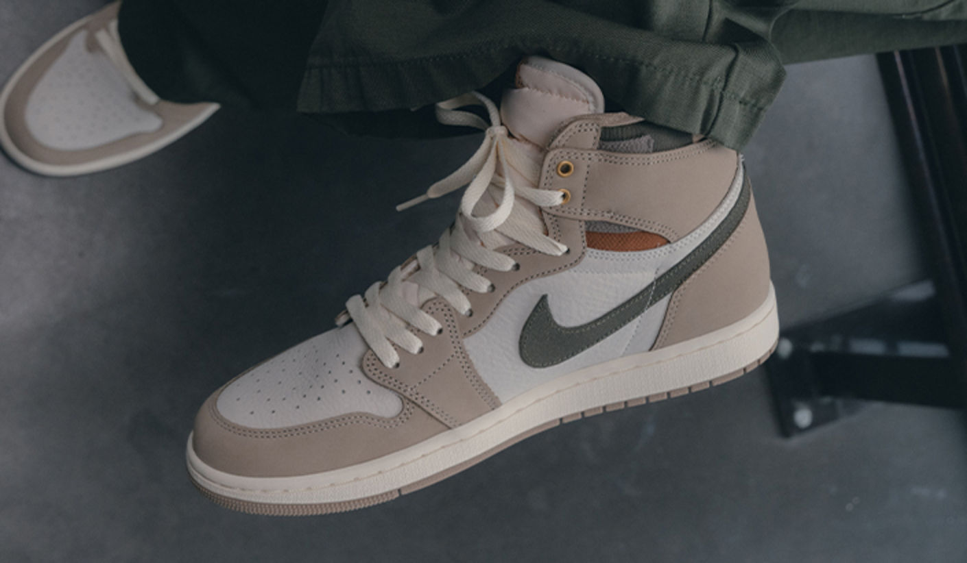 Close-up of an Air Jordan 1 Retro High OG “Khaki” sneaker in beige and white with a green Swoosh, worn with loose dark pants, set against a dark indoor floor.