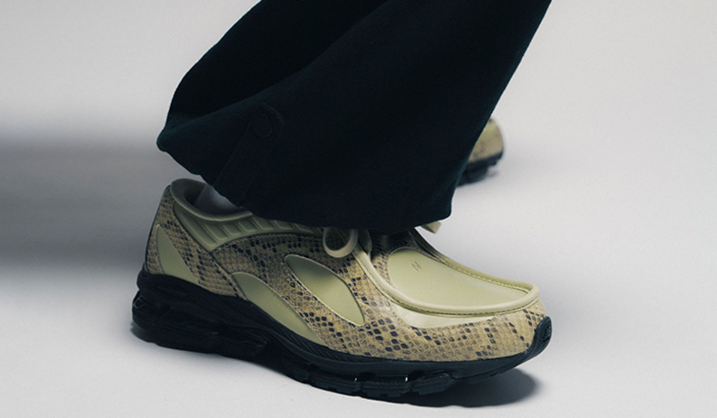 Close-up of New Balance U18907P8 sneakers in light green with textured, snake-inspired overlays and a black sole, worn with loose black pants against a minimal studio background.