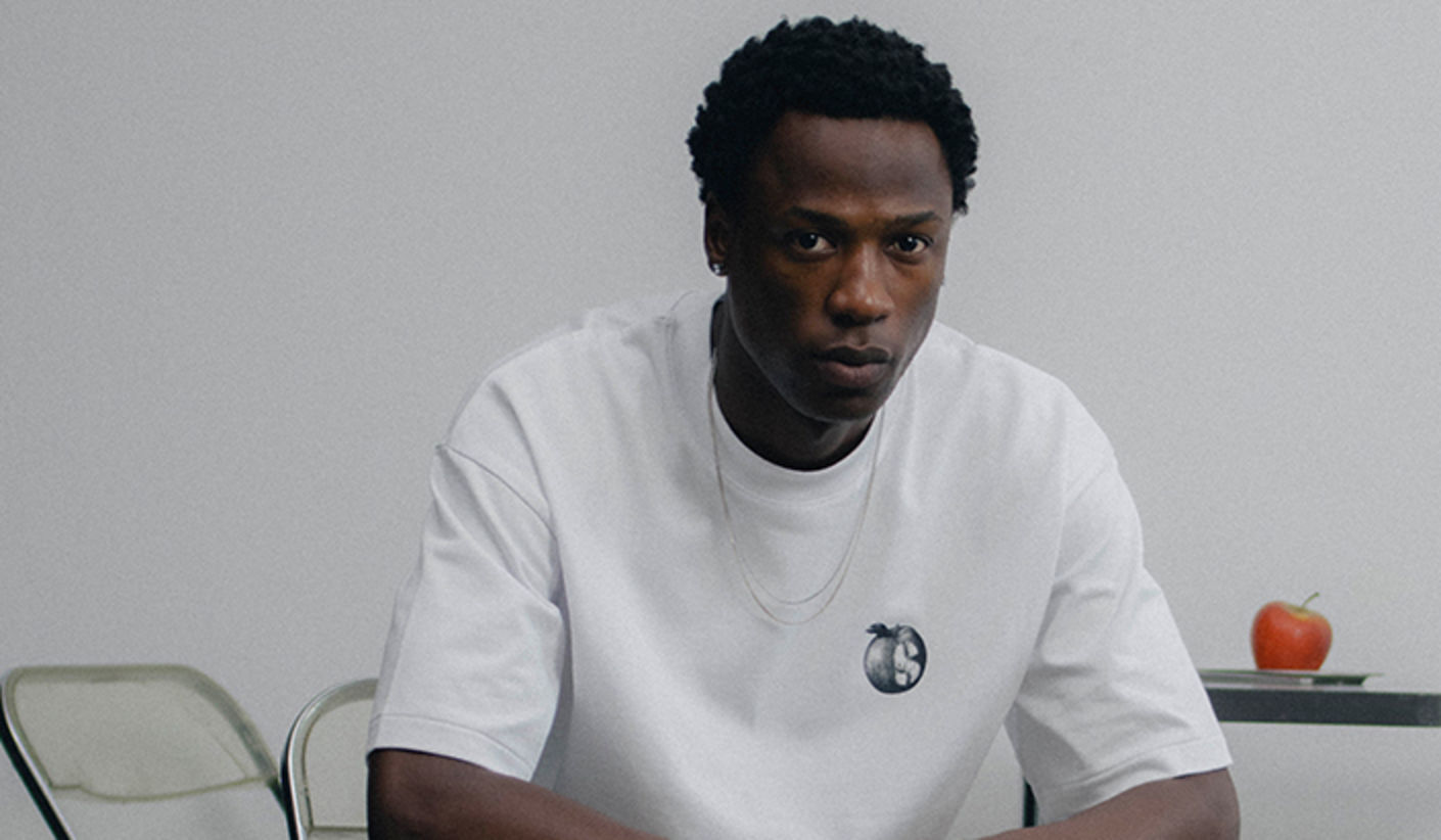 Front view of a person wearing the solebox Apple Tee from the Ordinary Objects collection, featuring a small apple graphic on a white T-shirt, styled in a minimalist interior setting.