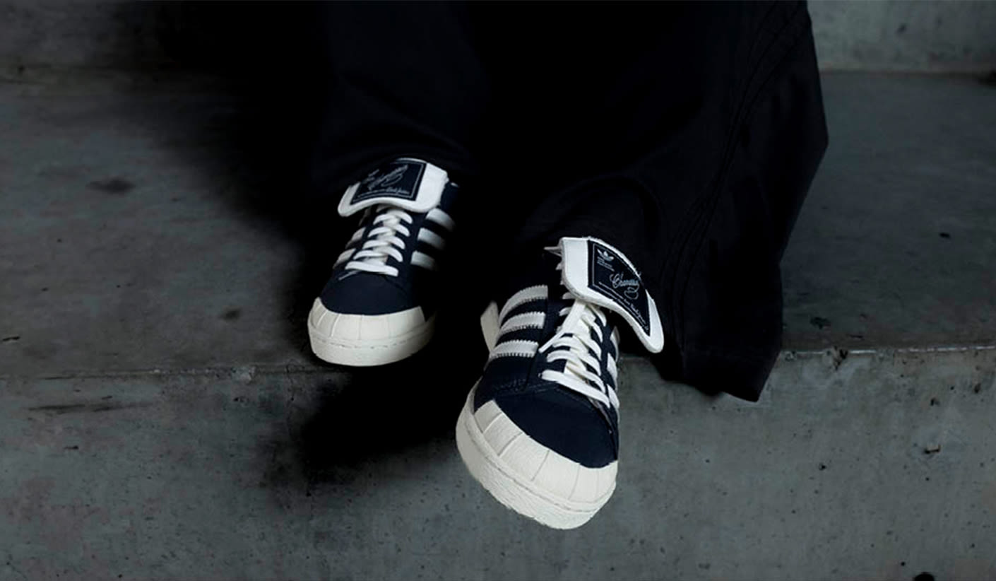 A close-up of a person sitting on concrete steps, wearing dark navy-and-white Adidas sneakers with oversized tongues and white laces. Only the shoes and the lower part of the pants are visible.