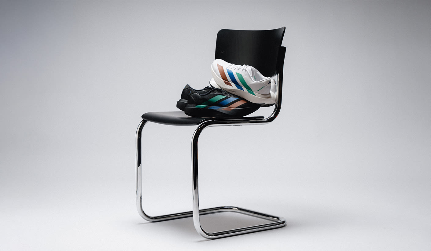 "Two pairs of Adidas sneakers placed on a modern black chair with chrome legs. One pair is white with multicolor stripes in blue, green, and orange, while the other is black with similar colored stripes. The setting is minimalistic with a neutral gray background."