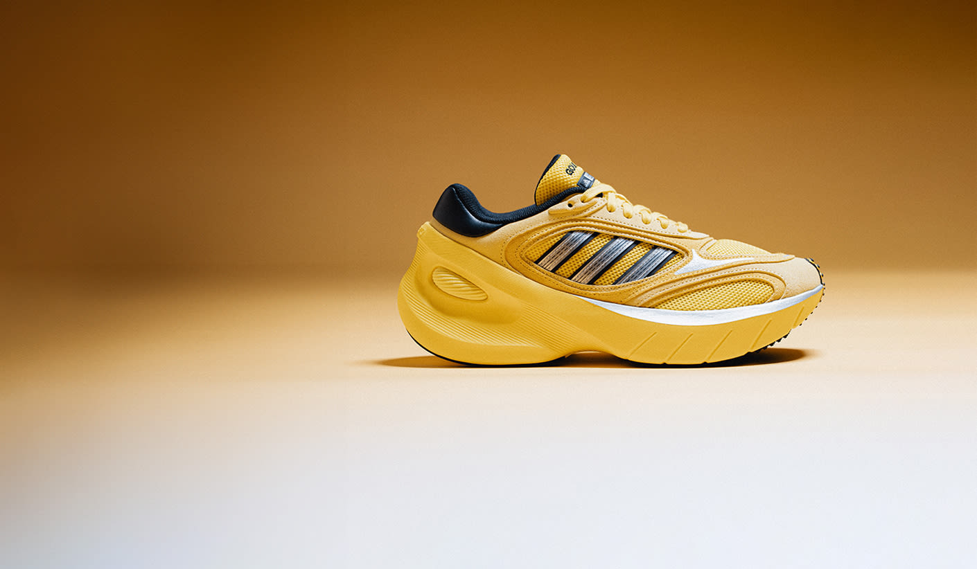 Side view of a bright yellow Adidas sneaker with a chunky sole and black and silver stripes, placed on a gradient yellow background