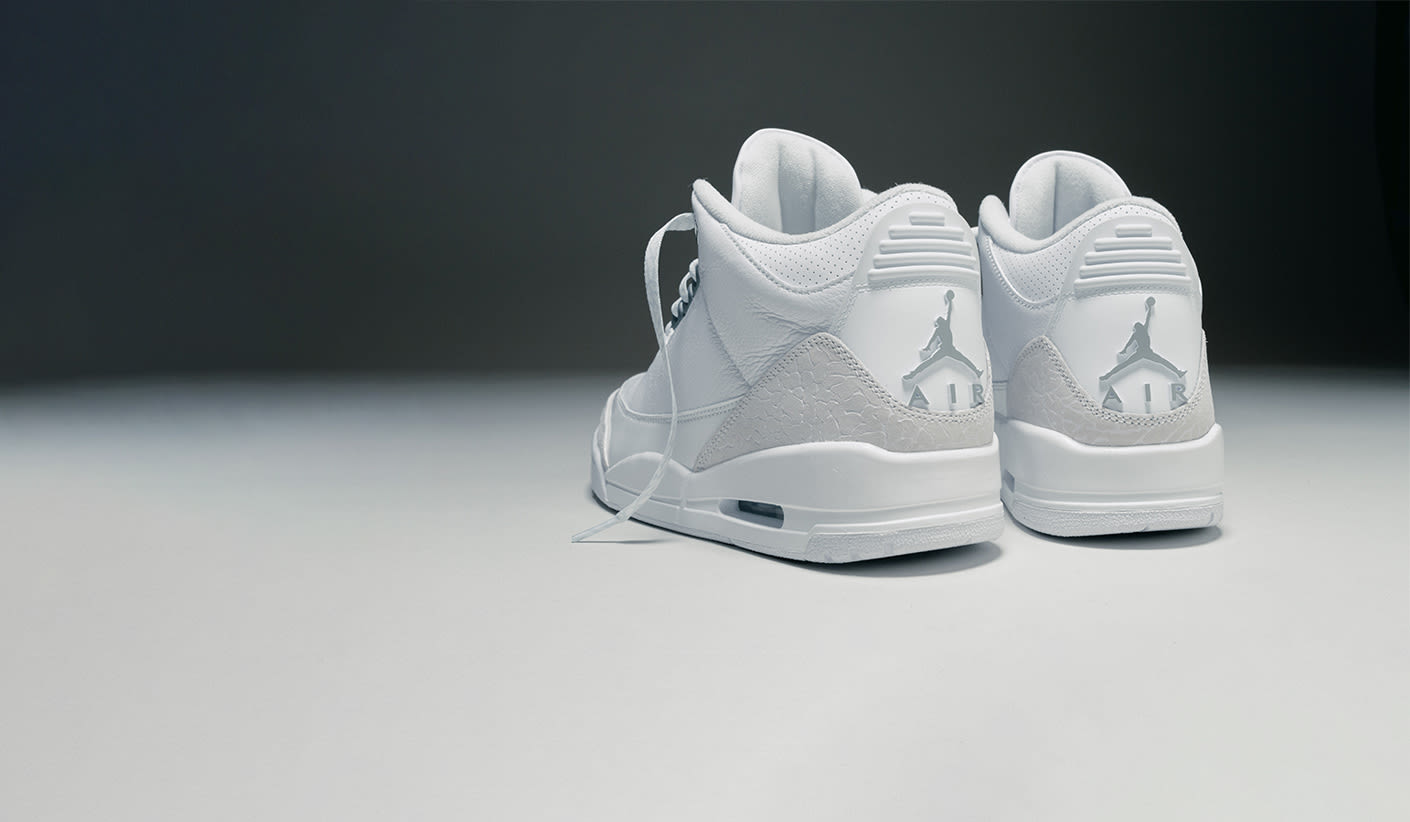 A pair of white Air Jordan 3 sneakers viewed from the back, showing the iconic Jumpman logo and the word "AIR" on the heel, placed on a minimalist grey and white background.