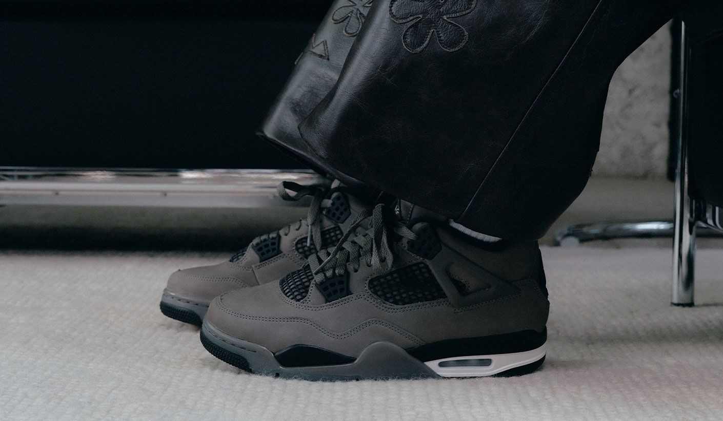 "A close-up side view of someone wearing dark grey Air Jordan 4 sneakers with black and white accents, paired with black leather pants with floral embroidery."
