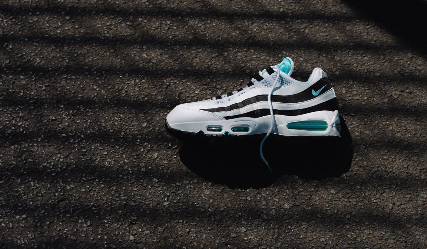 A Nike Air Max 95 sneaker in white, black, and teal colors lying on an asphalt surface, partially covered by striped shadows.