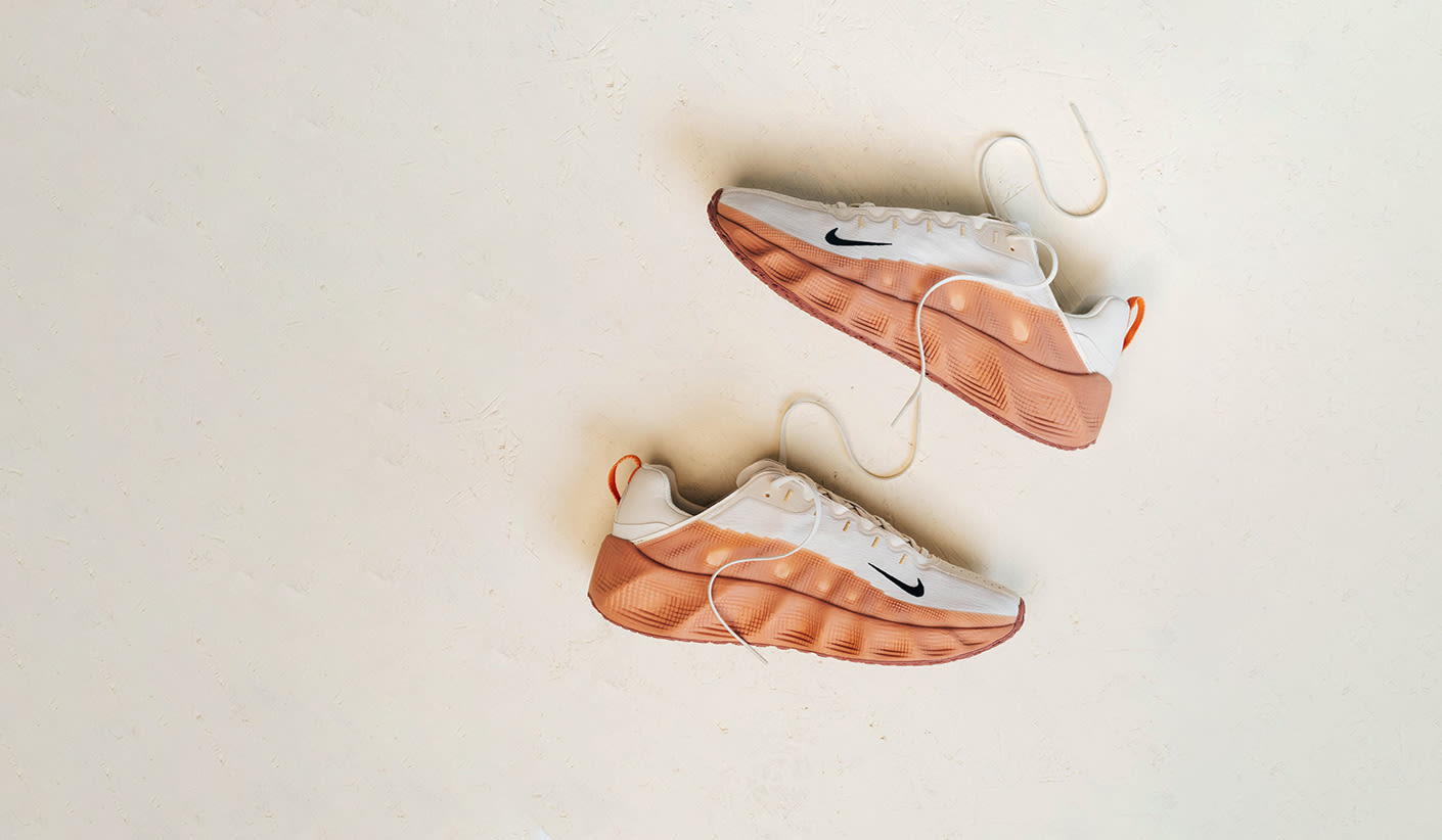 The image features a pair of Nike Ava Rovers, with a white upper and gradient orange sole, set on a clean, neutral surface. The laces are untied, giving a casual, modern look.
