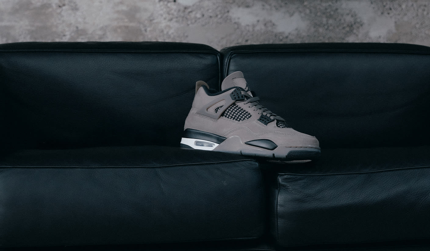 A single taupe-grey sneaker is placed on a black leather couch, set against a rough concrete wall. The shoe resembles an Air Jordan 4 with visible Air cushioning, black and white accents, and mesh side panels.