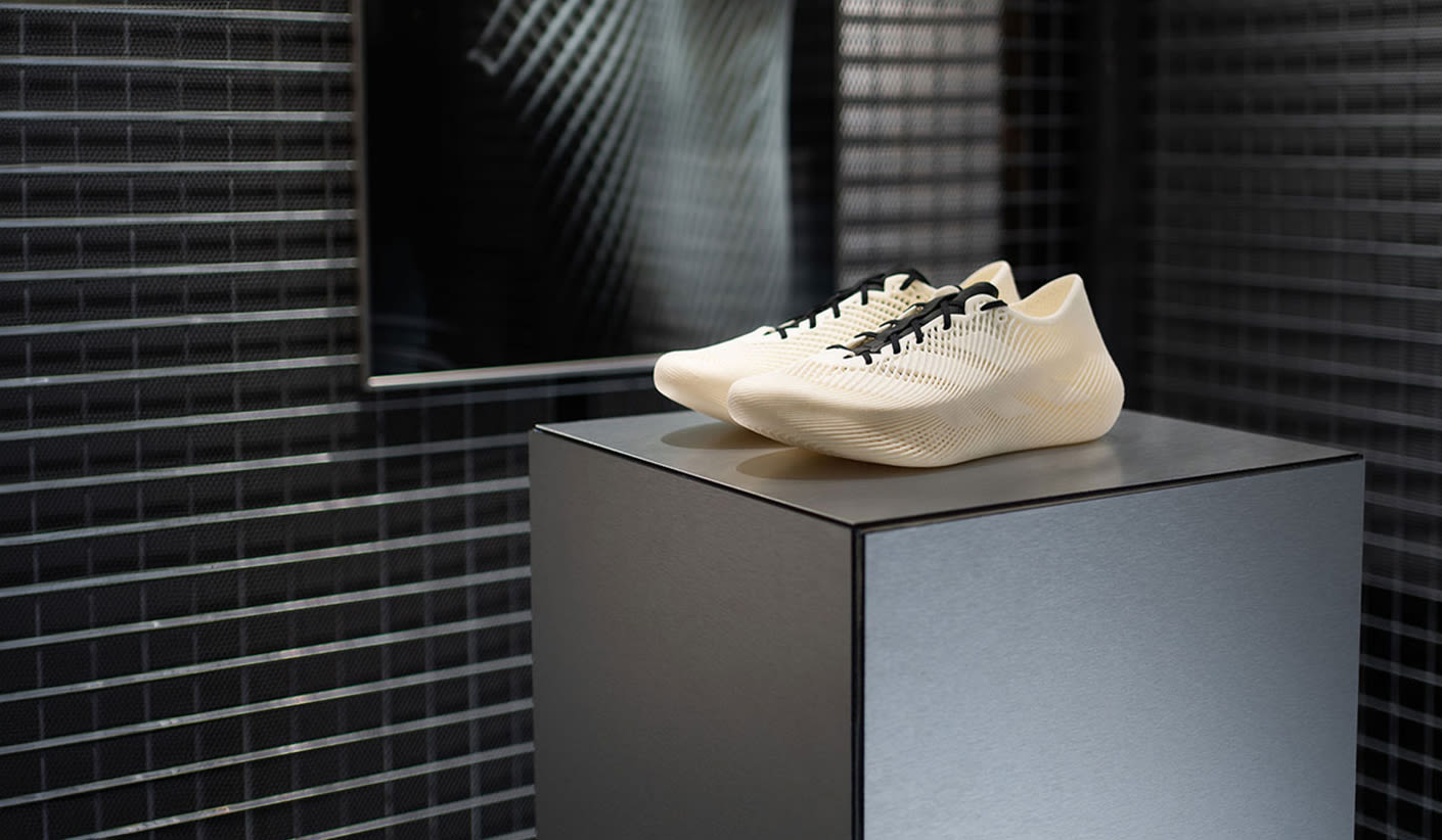 A pair of cream-colored athletic shoes with black laces sits on a dark gray pedestal. The shoes have a textured, possibly knitted or woven, upper and a thick, sculptural sole. In the background, there's a wall with small, dark, possibly black, square tiles, creating a grid pattern. A mirror or framed artwork with a striped, abstract design is visible on the wall behind the shoes.