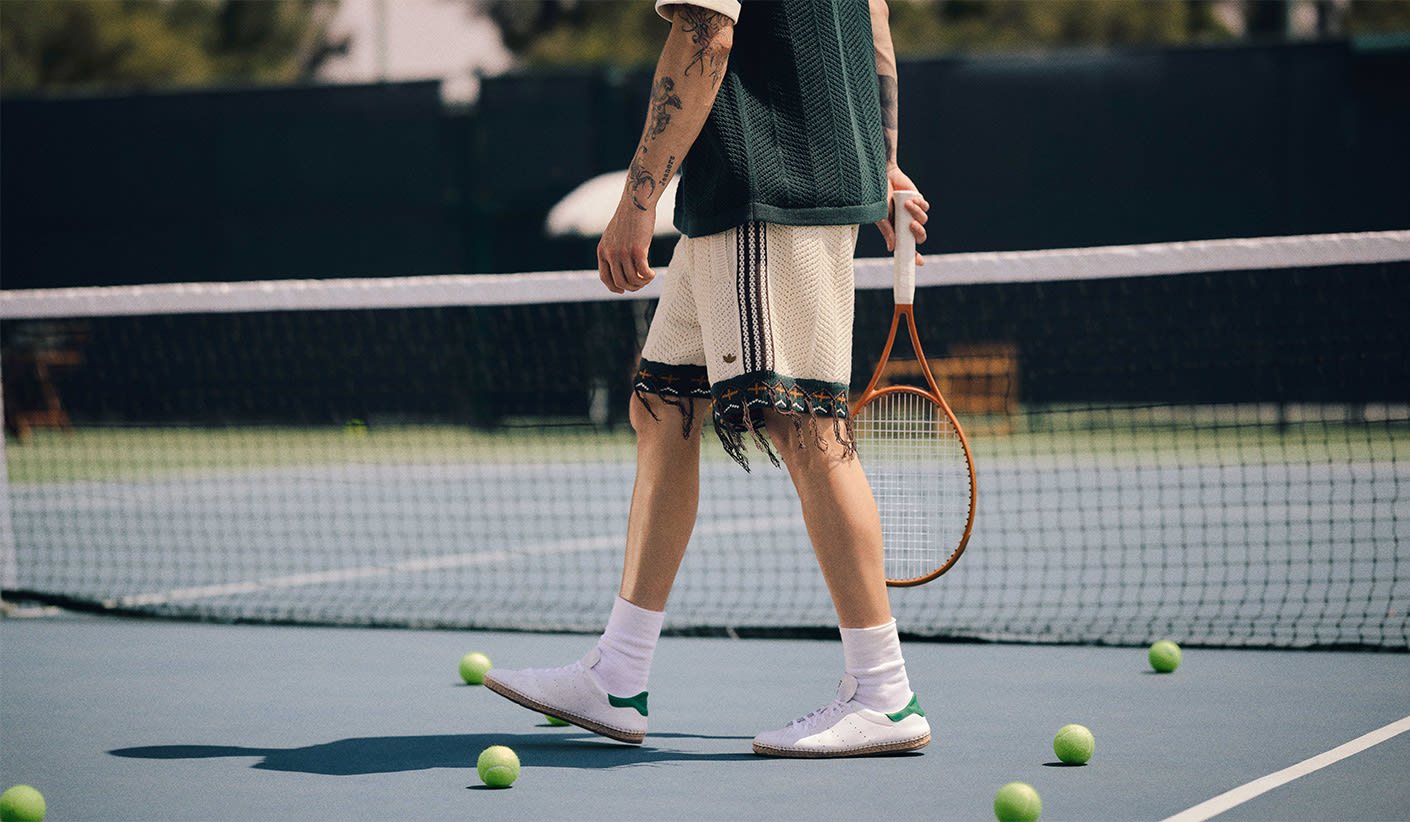 A person holding a tennis racket walks across a tennis court surrounded by tennis balls, wearing a knitted outfit with fringe details and white sneakers with green accents.