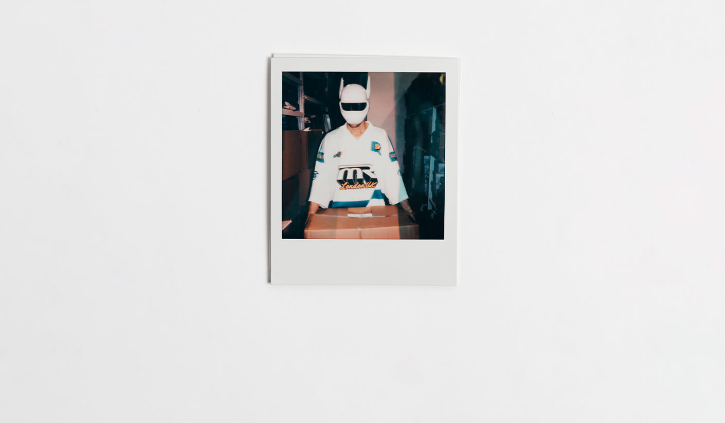 A Polaroid-style photo of a person wearing a futuristic white mask and a white hockey jersey with "London UK" and other graphic patches, holding a cardboard box in a storage room filled with shelves and boxes. The person is wearing Cro x Zellerfeld shoes.