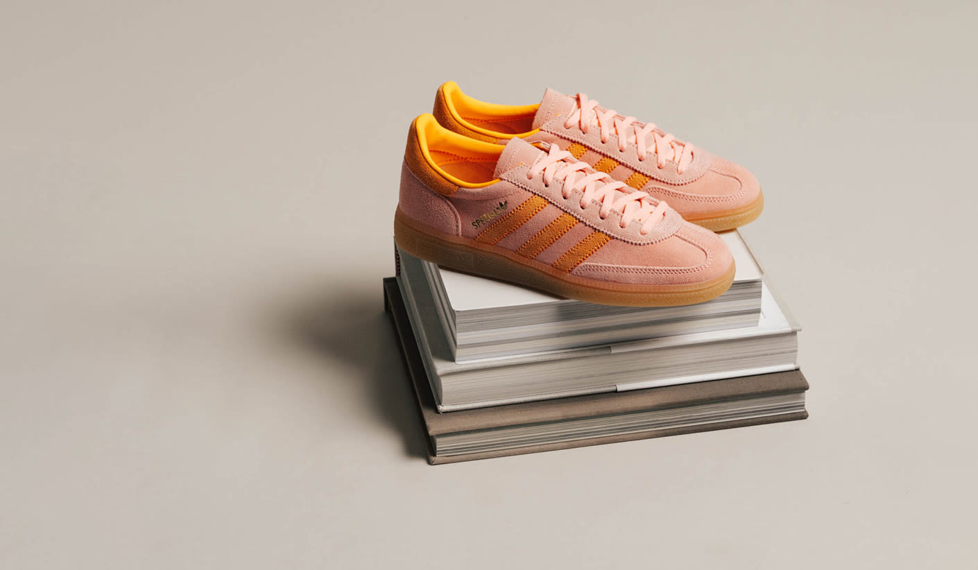 A pair of Adidas Handball Spezial sneakers in soft pink suede with bright orange stripes and inner lining, placed on top of a stack of books against a neutral beige background.