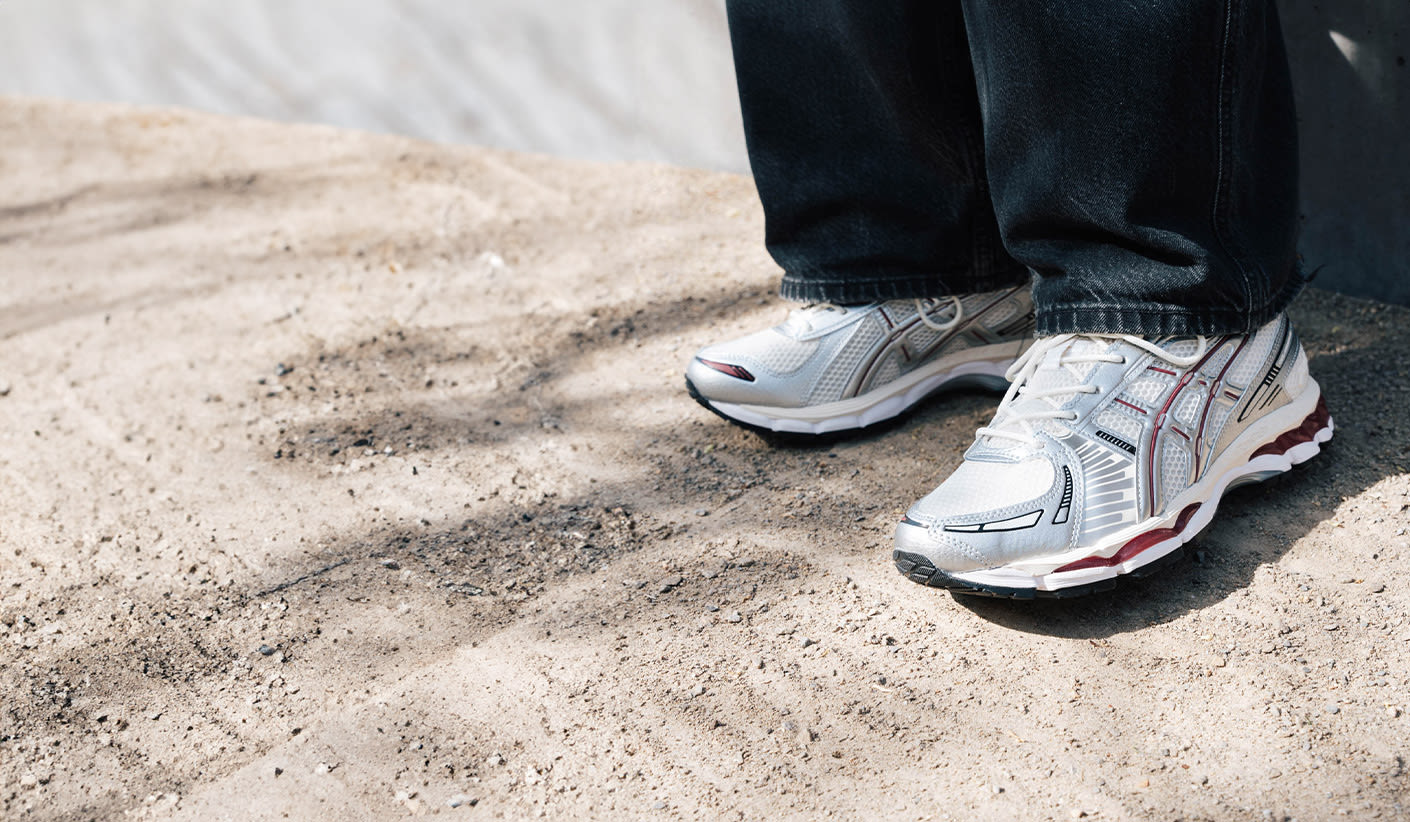 "Person wearing silver Asics Gel-Kayano 14 sneakers with red and black details, standing on sandy ground."