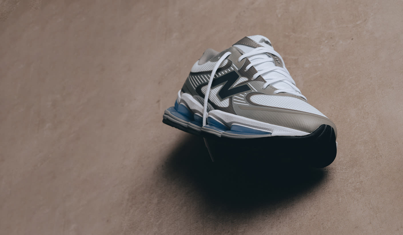 A single New Balance sneaker is showcased on a neutral-toned background, featuring white mesh, grey overlays, and blue cushioning details.