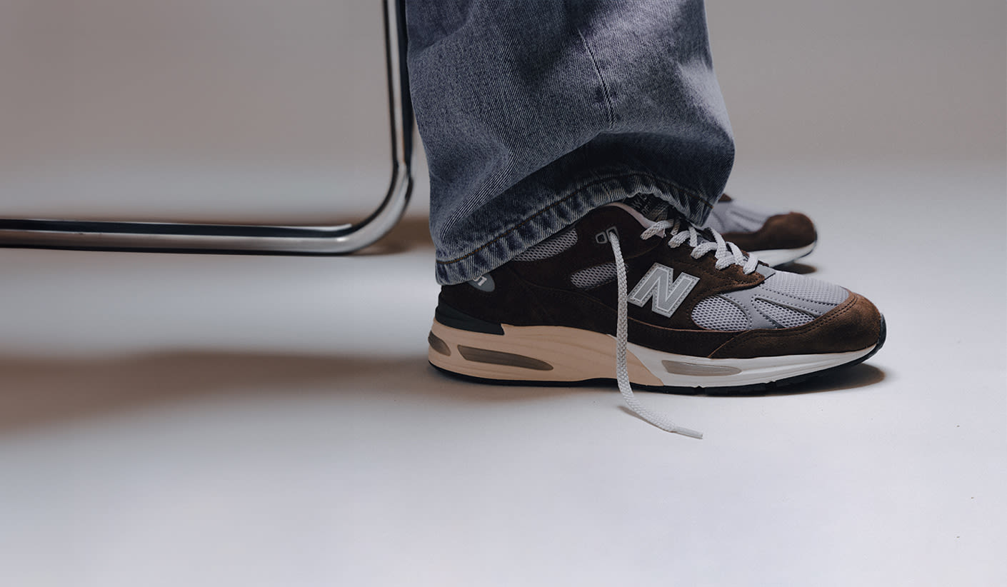 Person wearing brown and grey New Balance 991 sneakers with wide-leg jeans, sitting next to a metal chair on a light grey floor"