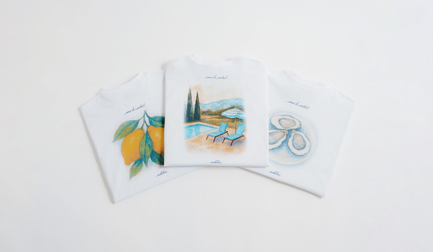 Three folded white T-shirts laid out side by side, each featuring a colorful illustration: lemons on the left, a poolside scene with chairs and umbrella in the center, and oysters on the right.