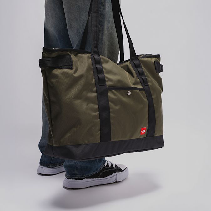 Olive green tote bag with black straps and base panel, carried with loose-fit jeans and black low-top sneakers.