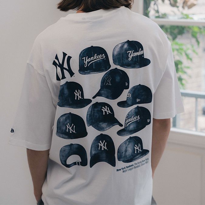 Back view of a person wearing a white New Era MLB New York Yankees graphic T-shirt featuring multiple Yankees cap illustrations and the NY logo, standing near a window.