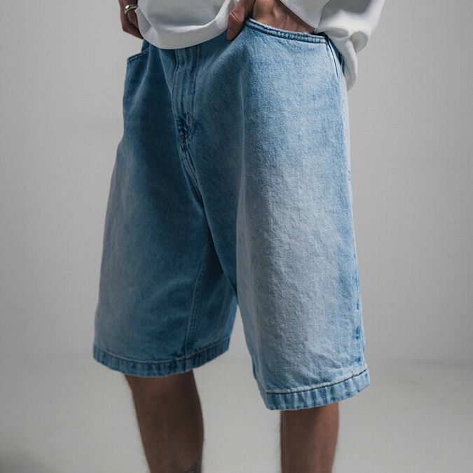 Mid-length view of a person wearing light blue denim shorts and a white long-sleeve top, with hands in pockets, styled against a plain neutral background.