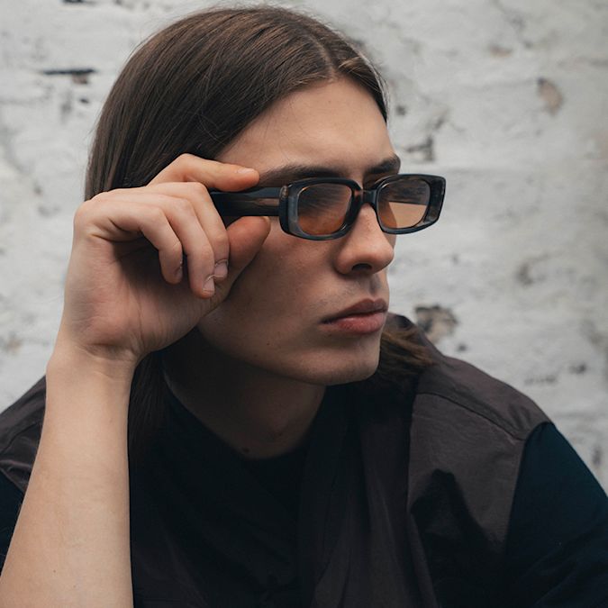 Close-up of a person adjusting brown rectangular “Legacy” sunglasses with tinted lenses, wearing a dark top against a textured light wall background.