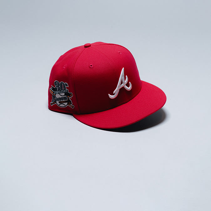 A red baseball cap is displayed on a light gray surface. The cap features a white, stylized "A" logo on the front. On the right side of the cap, there is a black and white patch with "30th Anniversary" text and a baseball graphic.
