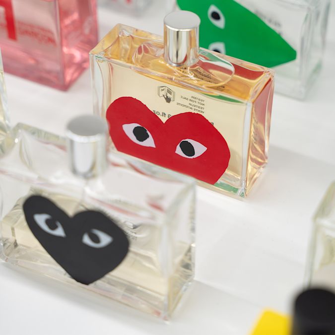 The image features a collection of perfume bottles, each adorned with distinctive heart-shaped designs featuring eyes. The colorful graphics on the bottles seem to evoke a playful and artistic style, likely inspired by the work of the fashion brand Comme des Garçons, known for its avant-garde and bold aesthetic. The eyes on the hearts could be a reference to the brand's signature motif, emphasizing a unique and expressive visual identity for their perfumes.