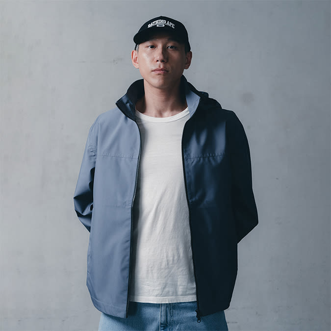 A person wearing a black baseball cap with white lettering, a light blue zip-up jacket over a white t-shirt, and light-wash jeans, stands against a plain concrete wall.