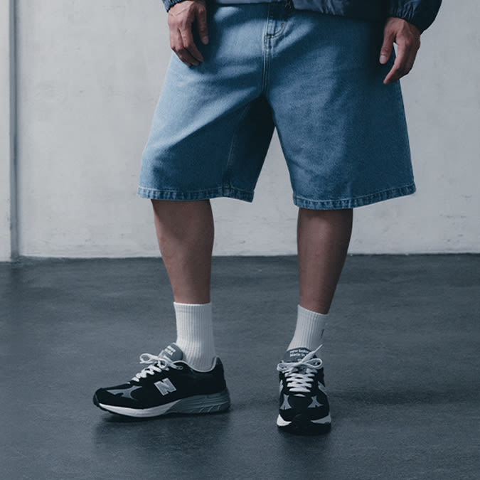 A person stands wearing light-wash denim shorts, a dark blue jacket, white socks, and a pair of dark New Balance sneakers with white laces. The background is a plain concrete wall and floor.