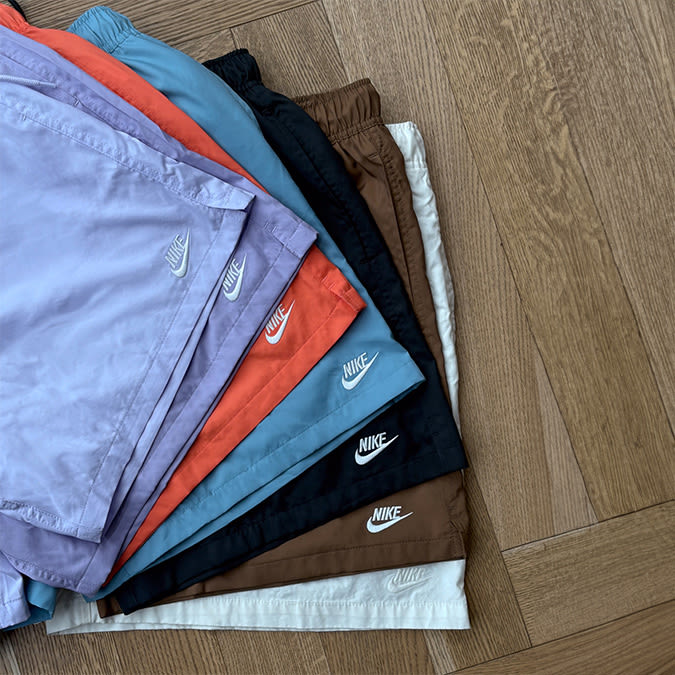 A neatly arranged stack of Nike shorts in various colors, including lavender, orange, light blue, black, brown, and off-white, displayed on a wooden floor with the Nike logo visible on each pair.