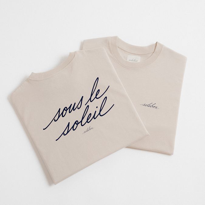 "Two folded beige T-shirts placed on a white surface. One features the handwritten text 'sous le soleil' in large navy letters and 'solebox' underneath. The other has a small 'solebox' logo on the chest."