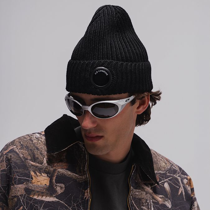 Person wearing a black rib-knit beanie with integrated lens detail, silver wraparound sunglasses, and a camouflage-pattern jacket against a light studio background.